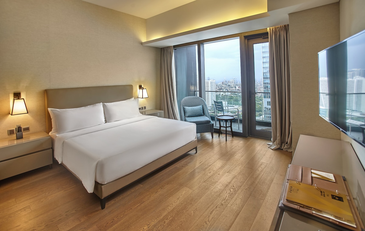 A spacious guest room features a king-sized bed, complemented by contemporary furnishings. Large glass doors provide access to a scenic city view, while a cozy chair sits beside a sleek bedside table. Natural light is welcomed through floor-to-ceiling curtains, enhancing the modern aesthetic.