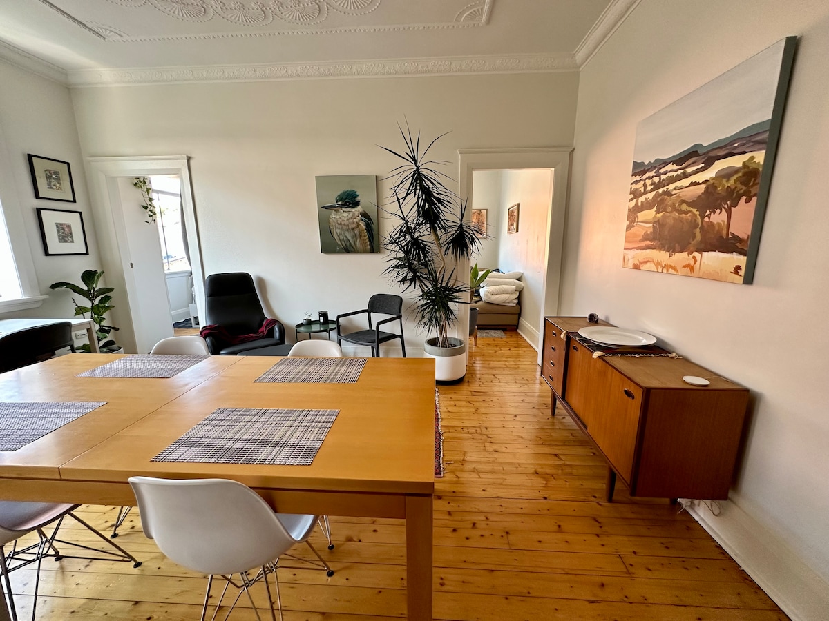 A spacious dining area features a large wooden table surrounded by several chairs. The room is enhanced by a tall indoor plant and artwork on the walls. Natural light enters from a window, illuminating the polished wooden flooring.