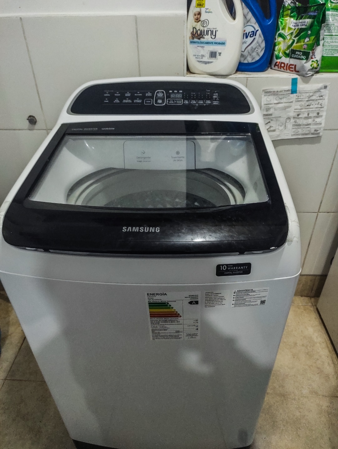 A front view of a modern washing machine is displayed in a bright laundry area. Detergent bottles are organized above the machine, and clear labels indicate various settings on the machine's control panel. The surrounding walls are tiled, contributing to a clean environment.