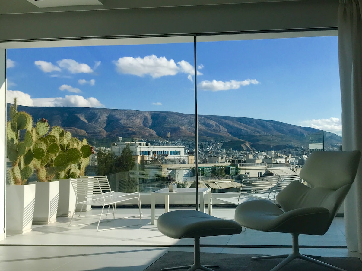 A panoramic view of Mount Hymettus is captured through large glass windows, showcasing the natural landscape against a blue sky. A modern seating arrangement, consisting of white chairs and a table, is positioned on the stylish floor, enhancing the open ambience of the living area.