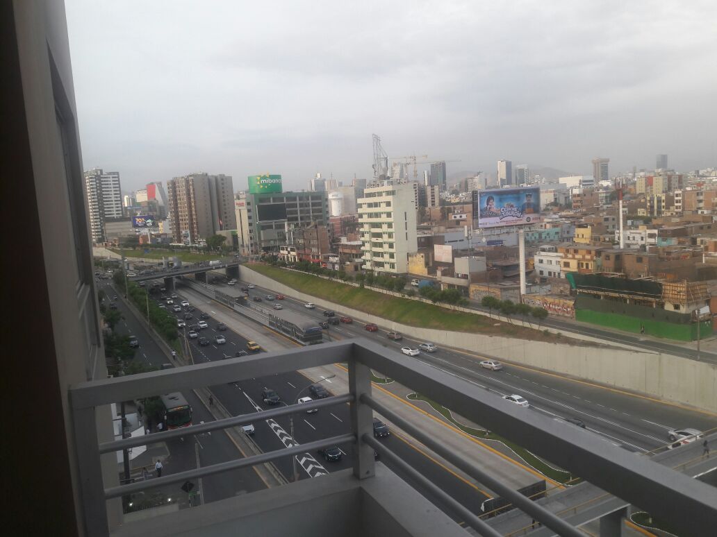 A view from the terrace shows a busy avenue, with vehicles traveling along multiple lanes. Buildings line the background, including tall commercial structures and billboards. The gray sky creates a muted atmosphere over the urban landscape.