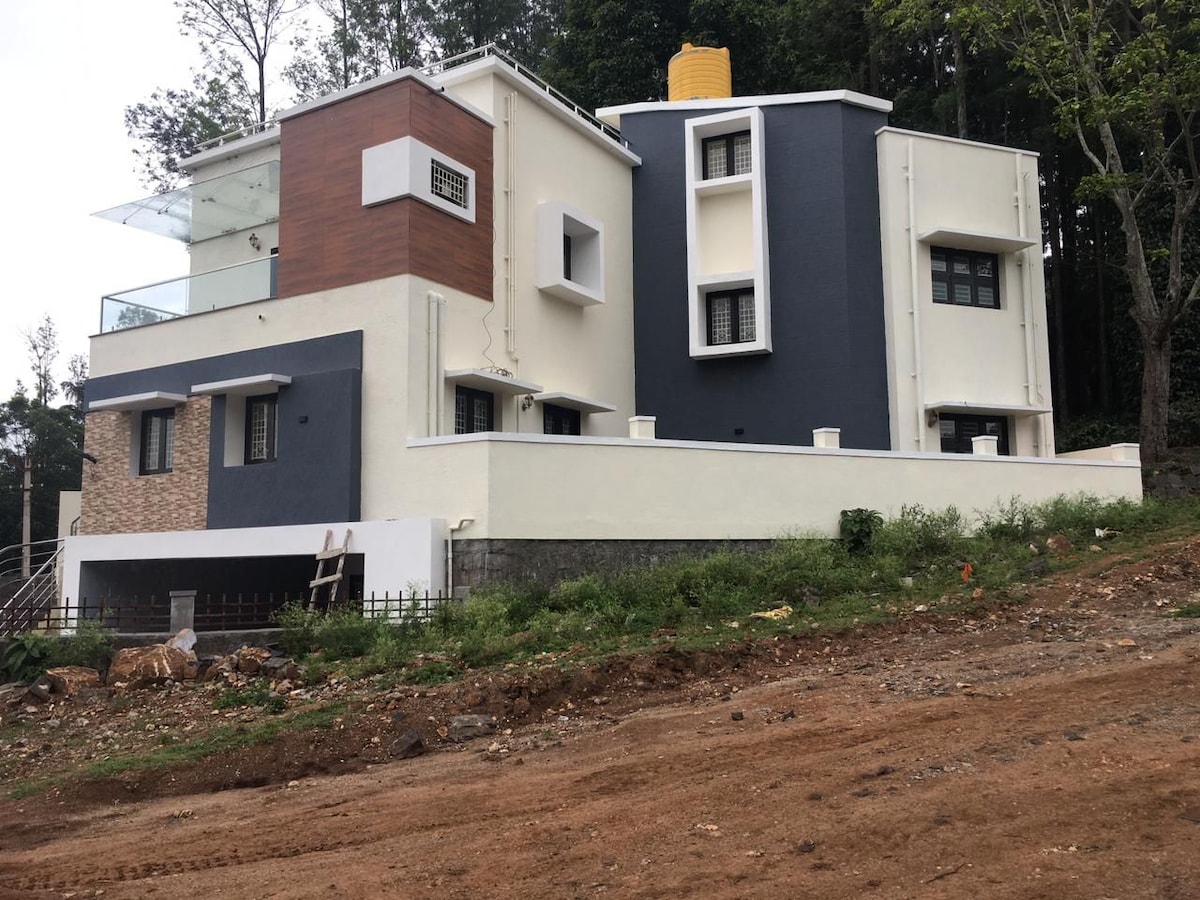A modern two-story house is set in a natural landscape, featuring a combination of light and dark exterior walls. Large windows are present on each level, and a balcony is observed on the upper floor, surrounded by green trees and a gravel pathway.