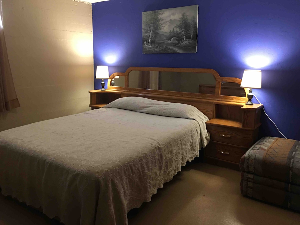 A cozy sleeping area features a spacious bed covered with a patterned blanket, complemented by a wooden headboard and matching side tables. Two lamps provide soft lighting, and a landscape painting adorns the blue wall above the bed. A small seating area is visible in the corner.