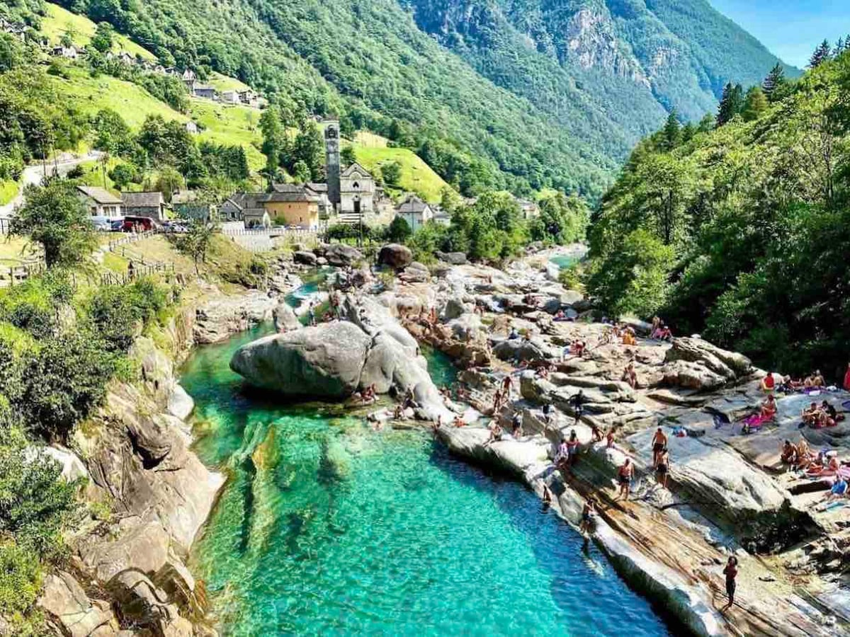 A scenic view of a river winding through a lush green valley, framed by rocky landscapes. People are seen enjoying the water along smooth, natural rock formations. A village with a bell tower is visible in the distance, nestled among the hills.