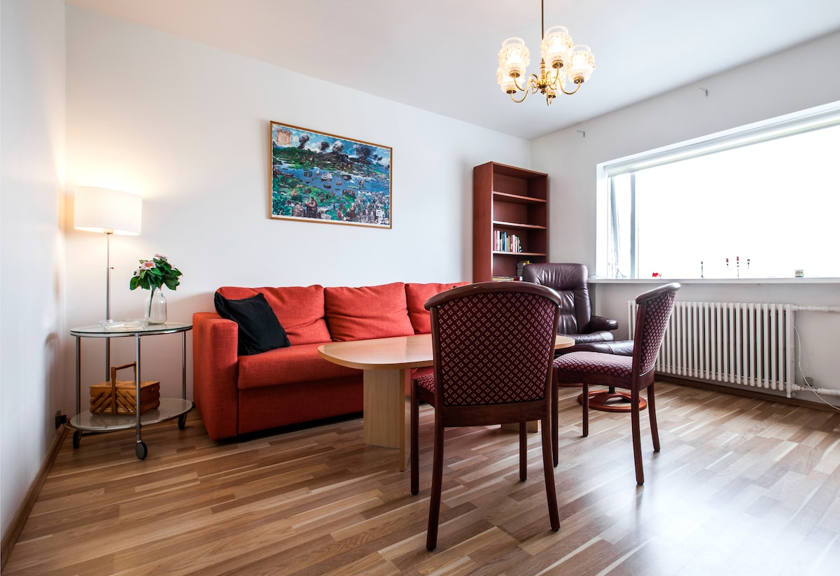 A bright living area features a red sofa and a circular wooden table with four chairs. A floor lamp stands beside a small potted plant on a glass side table. A large window offers natural light and the view of the outside, while a bookshelf is positioned against the wall.