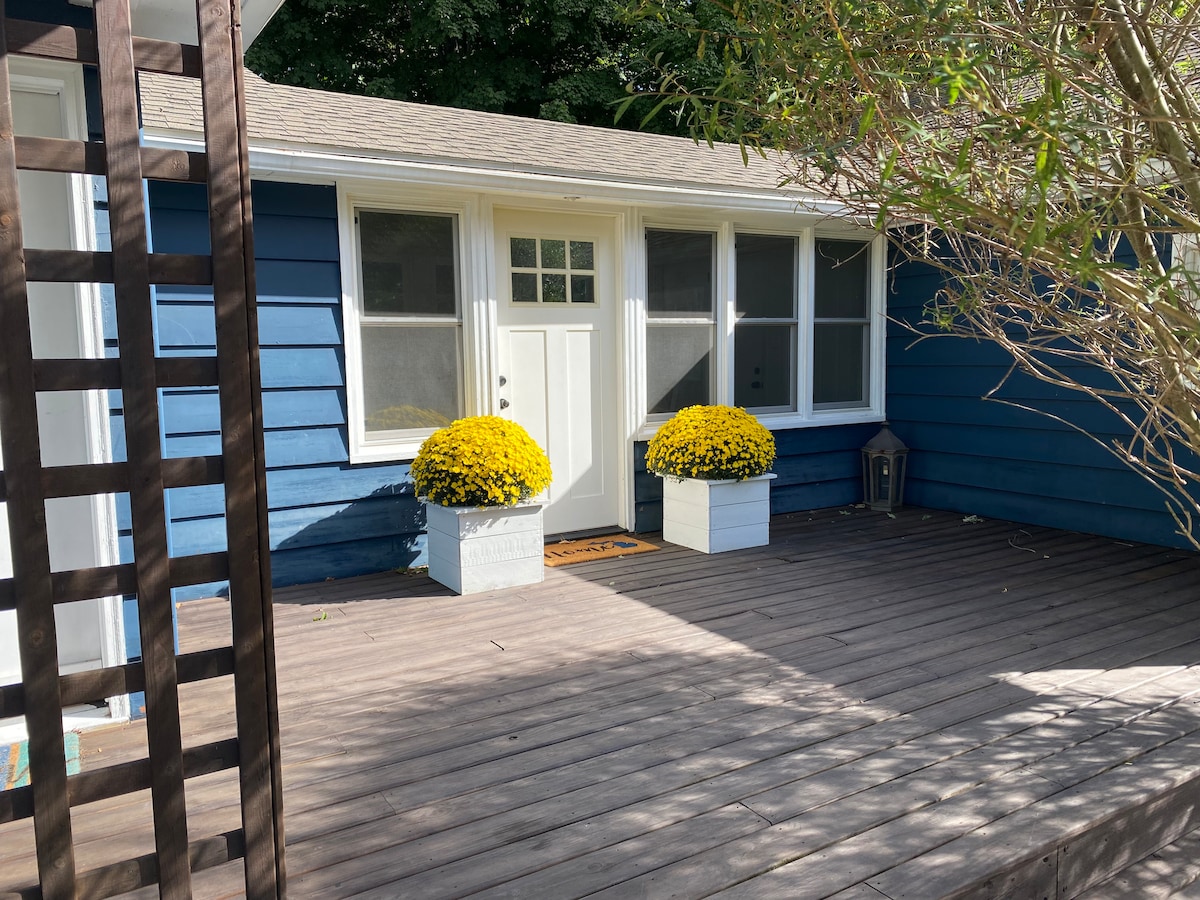 The entrance is framed by two large white planters filled with vibrant yellow mums, enhancing the welcoming ambiance. A wooden deck leads to a front door with a glass panel, surrounded by large windows that invite natural light into the home.