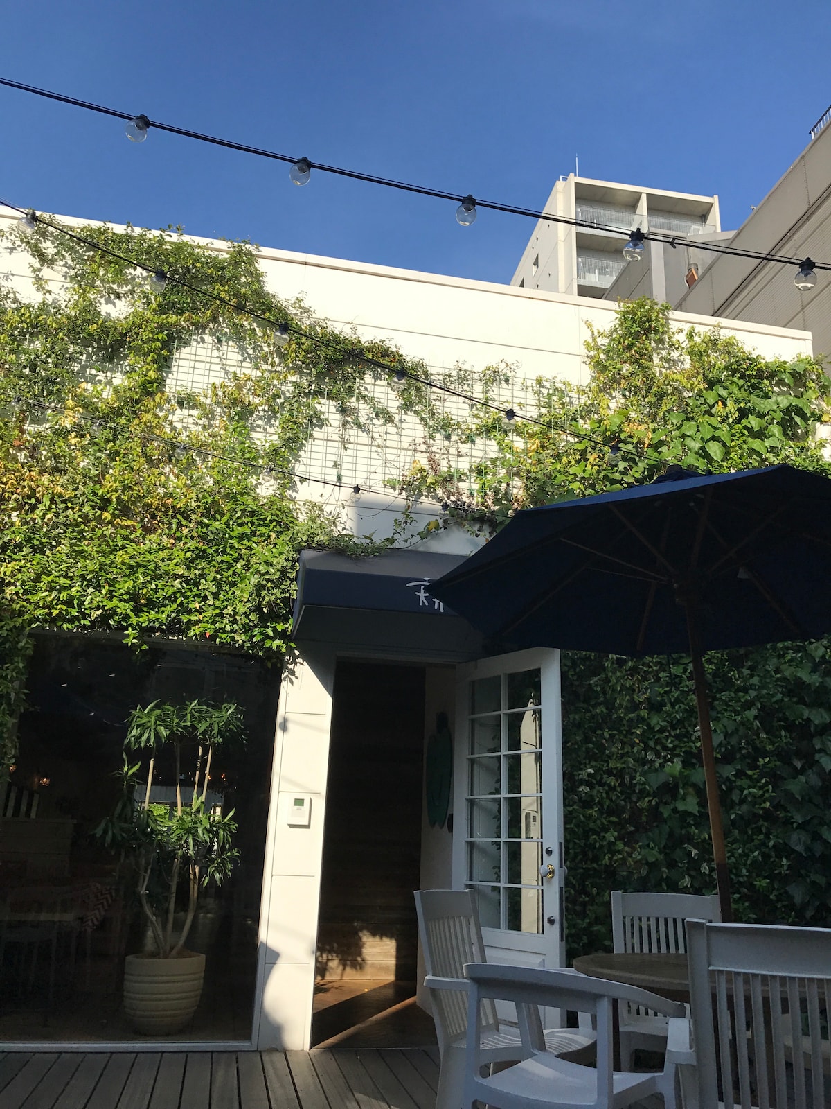The exterior of the property is adorned with lush green vines, creating a natural ambiance. A spacious outdoor area features white wooden furniture and a large umbrella. Clear blue skies enhance the inviting atmosphere of this tranquil space.