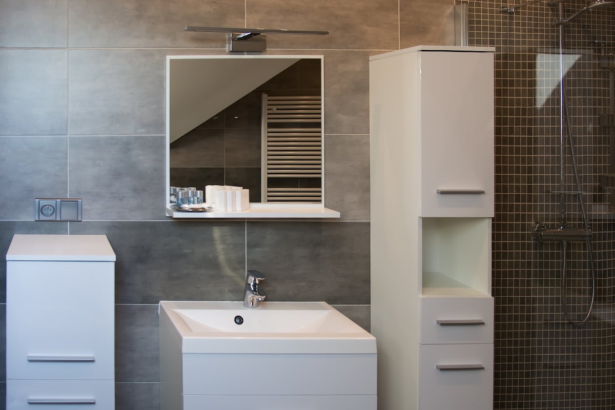 A modern bathroom features a sleek white vanity with a round sink and a large mirror above. A tall storage cabinet is positioned next to the vanity, and a tiled shower area is visible in the background, equipped with a chrome showerhead.
