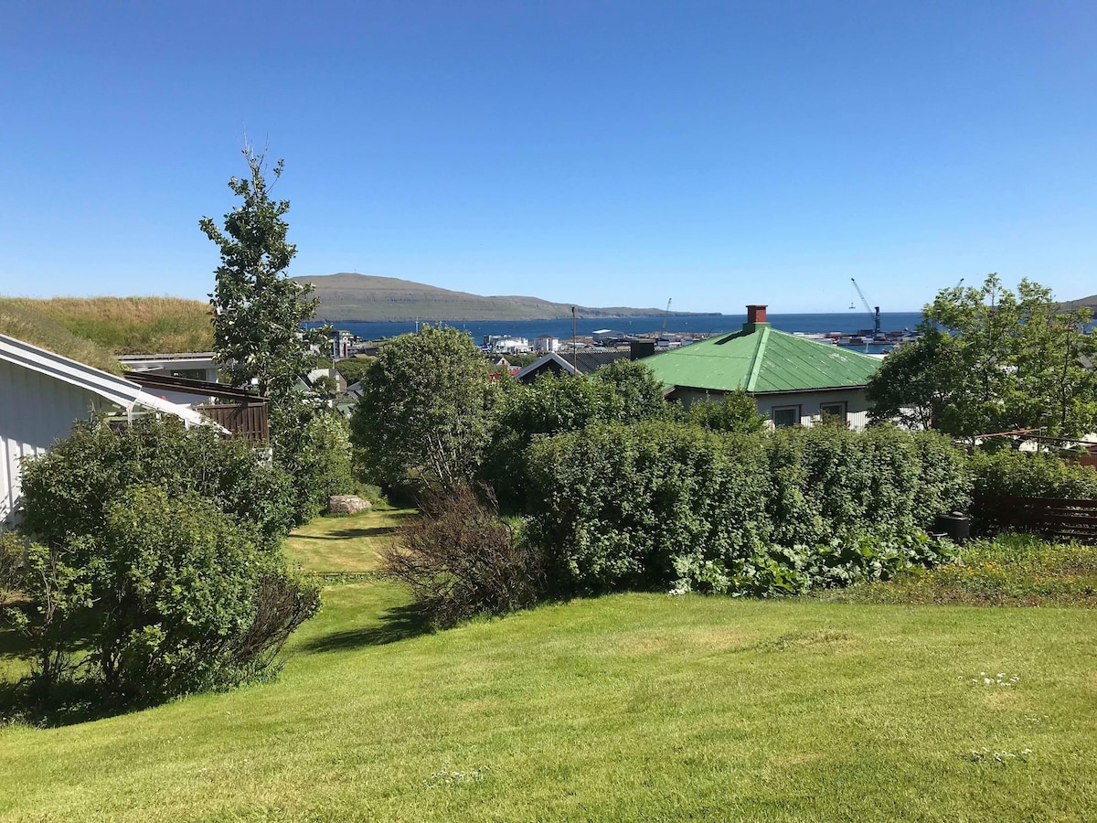 A view of a green lawn is featured, bordered by various shrubs and small trees. In the background, the blue waters can be seen beyond residential roofs, with distant hills and harbor activity visible under a clear sky.
