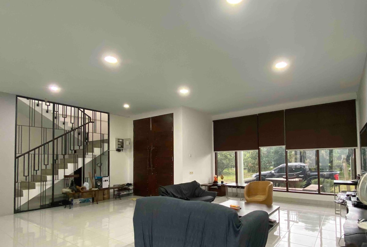 A spacious living area is framed by large windows, allowing for natural light and views of the outdoor surroundings. Comfortable seating is arranged around a central coffee table. A staircase is visible in the background, leading to an upper level.