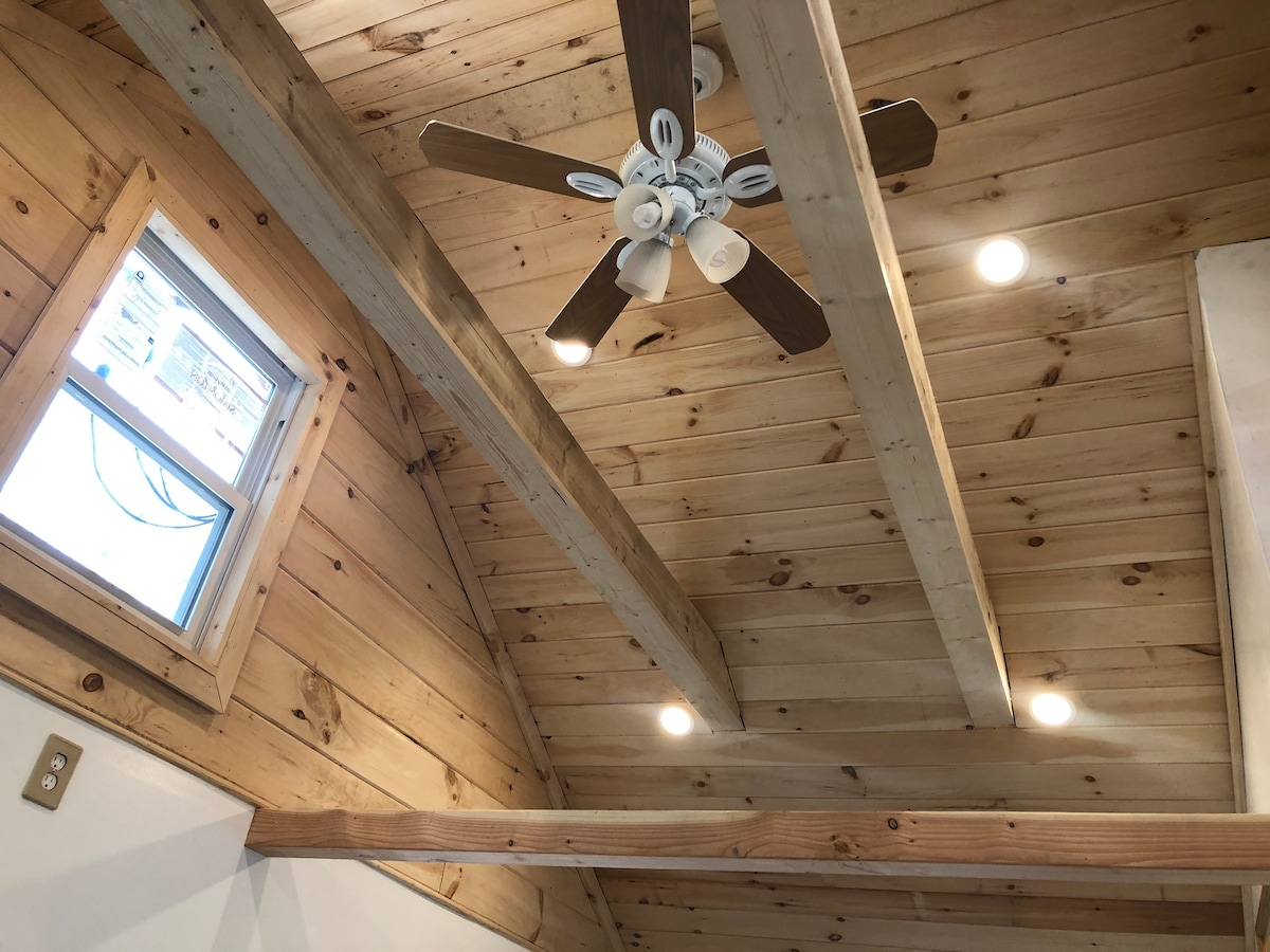 The ceiling displays wooden beams with a light wood finish, complemented by recessed lighting. A ceiling fan with five blades is positioned centrally, while a small window allows natural light to filter in, enhancing the warm aesthetic of the space.