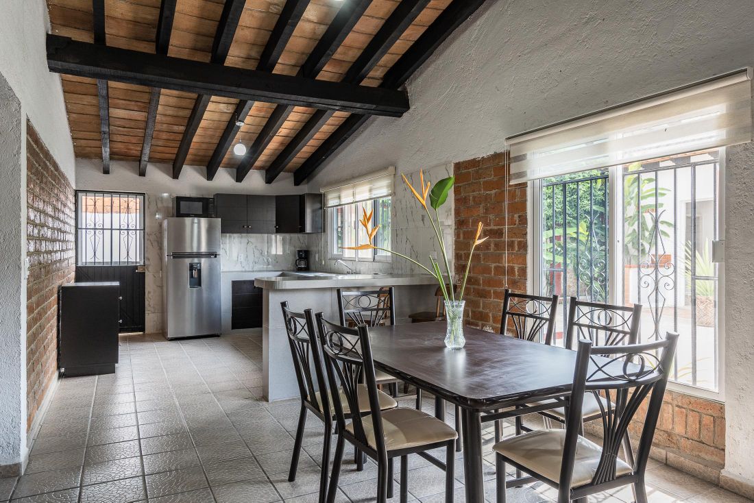 An open kitchen and dining area features a large table with eight chairs, adjacent to a kitchen counter. The space is illuminated by natural light through large windows, and wooden beams accent the ceiling, providing a rustic touch. A refrigerator and modern appliances are visible.