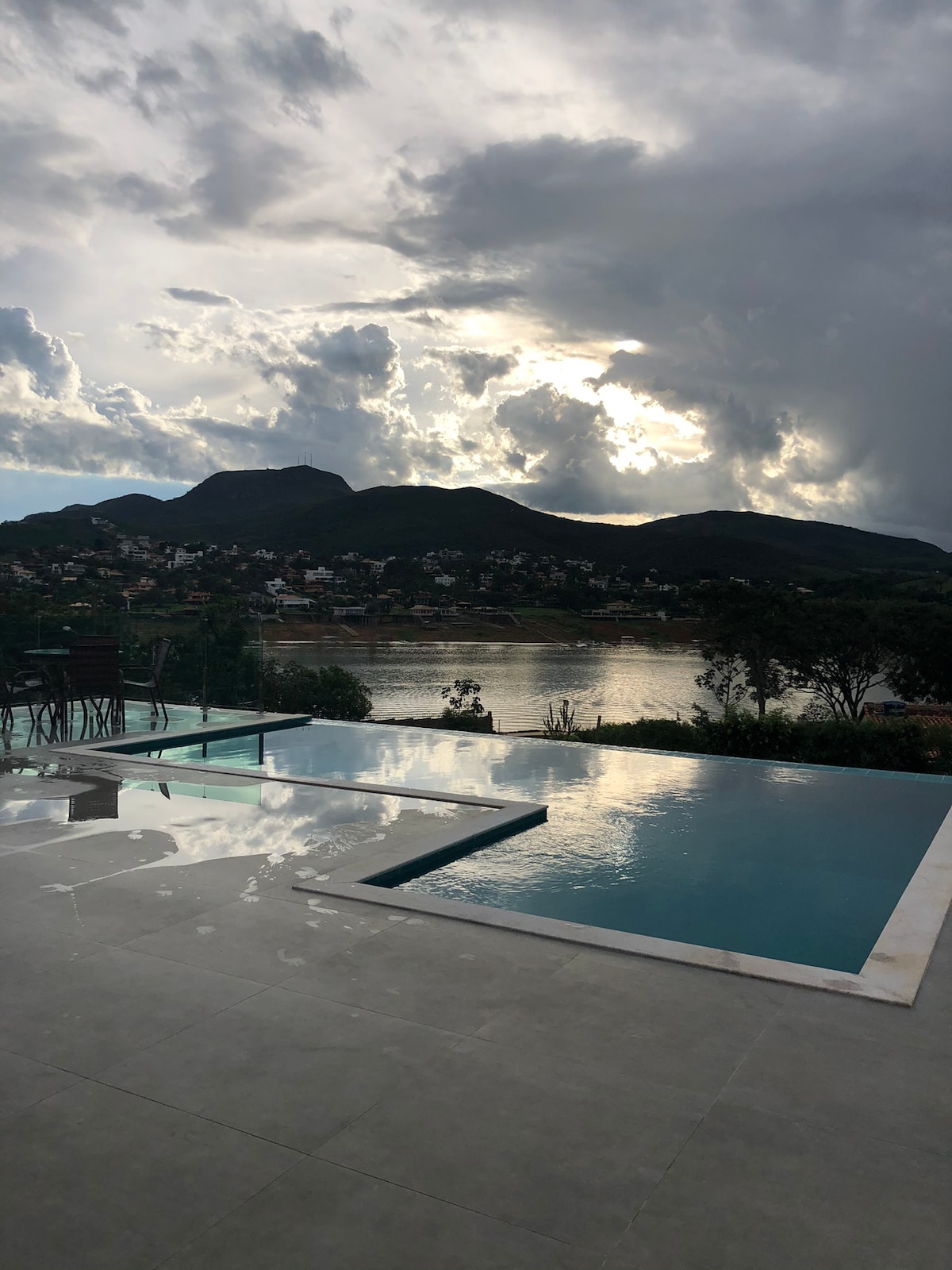 An outdoor area features a serene swimming pool with clear water reflecting the surrounding landscape. Soft clouds fill the sky above, while distant mountains provide a scenic backdrop. The calm lake is visible in the background, hinting at a tranquil atmosphere.