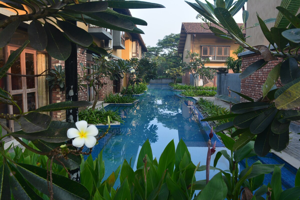A serene swimming pool is framed by lush green foliage and blooming flowers. Surrounding buildings provide a natural backdrop, creating a peaceful setting. Sunlight reflects off the water's surface, highlighting the inviting atmosphere of this private space.