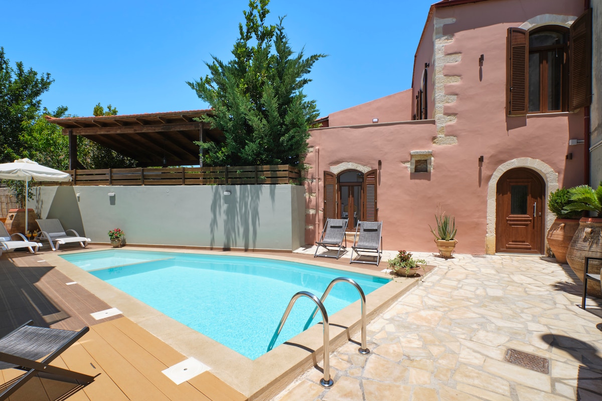 A stone villa features a private pool surrounded by a sun deck. Lounge chairs are positioned beside the clear water, and shaded areas are provided by an overhead structure. Lush greenery and decorative pots add to the serene outdoor ambiance.