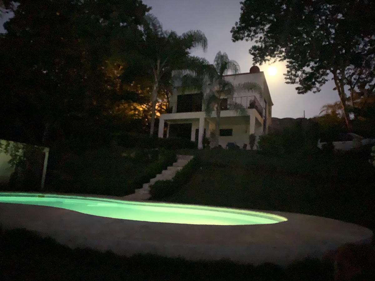 The image captures a tranquil scene of a glowing pool, framed by lush greenery and illuminated by moonlight. In the background, the two-story house is partially visible, with warm lights enhancing the serene atmosphere of the evening.