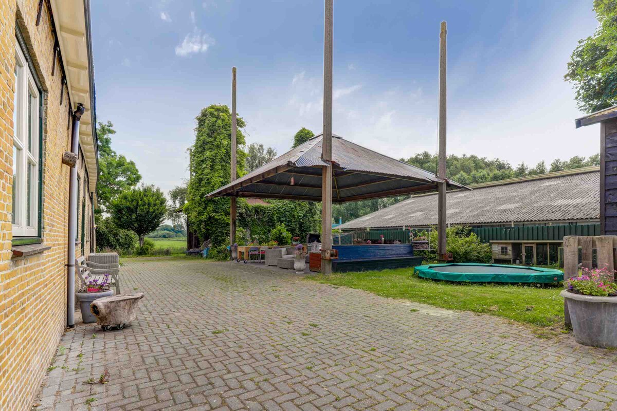 An outdoor space is presented with a large covered seating area, supported by wooden posts. A paved surface offers direct access to the surroundings, which include greenery and an inviting blue swimming pool. Furnishings provide a relaxing atmosphere for gatherings or leisure.