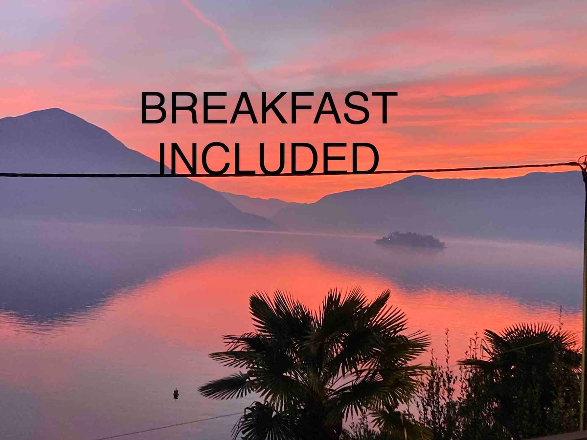 A serene sunset view features soft pink and orange hues reflected on the lake's surface. Silhouette of mountains is visible in the background, with a palm tree in the foreground framing the scene.