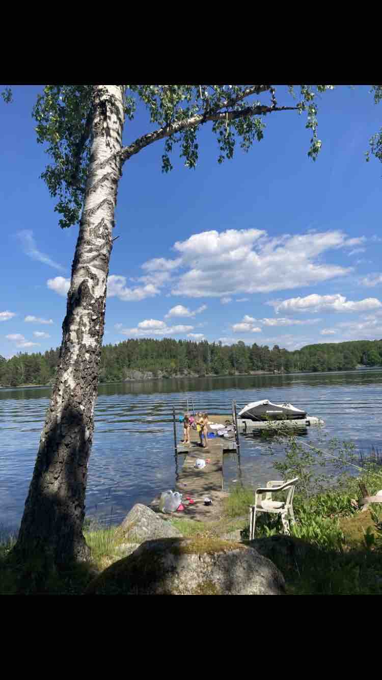 A dock extends over a calm lake, surrounded by greenery and a sandy shoreline. A small boat is moored at the dock, while people can be seen engaging in leisure activities. Sunlight reflects off the water under a bright blue sky with scattered clouds.