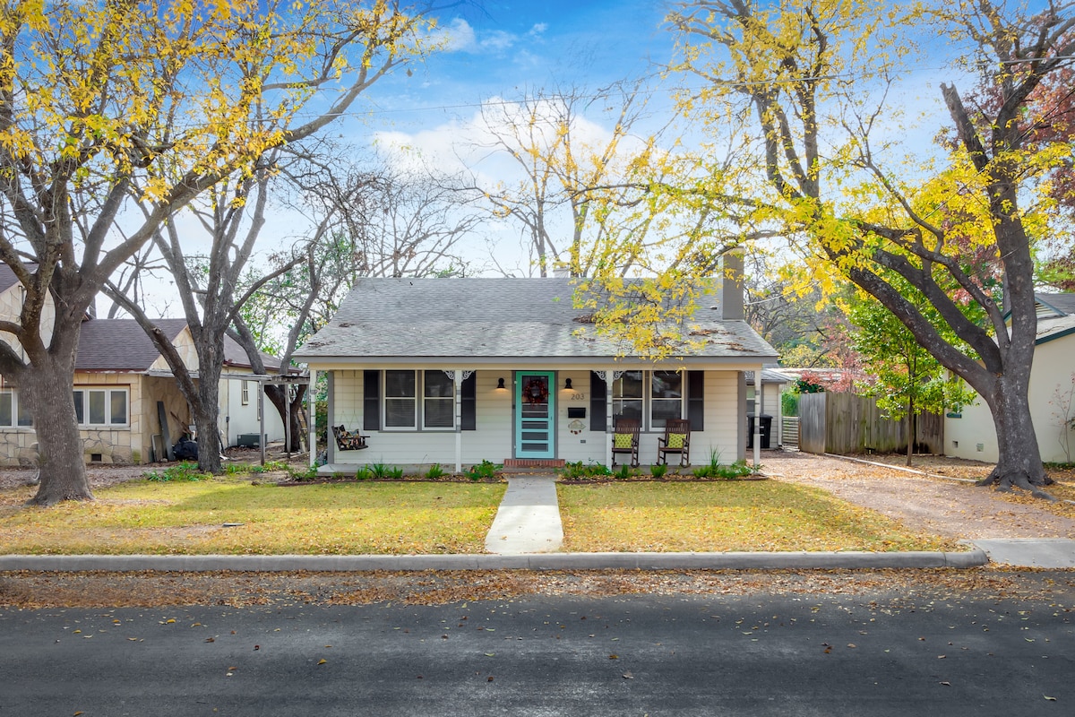 A charming single-story cottage is framed by tall trees with yellow leaves. A welcoming front porch features a blue door and two dark chairs, with a well-maintained lawn leading up to the entrance. The surrounding neighborhood appears peaceful and inviting.