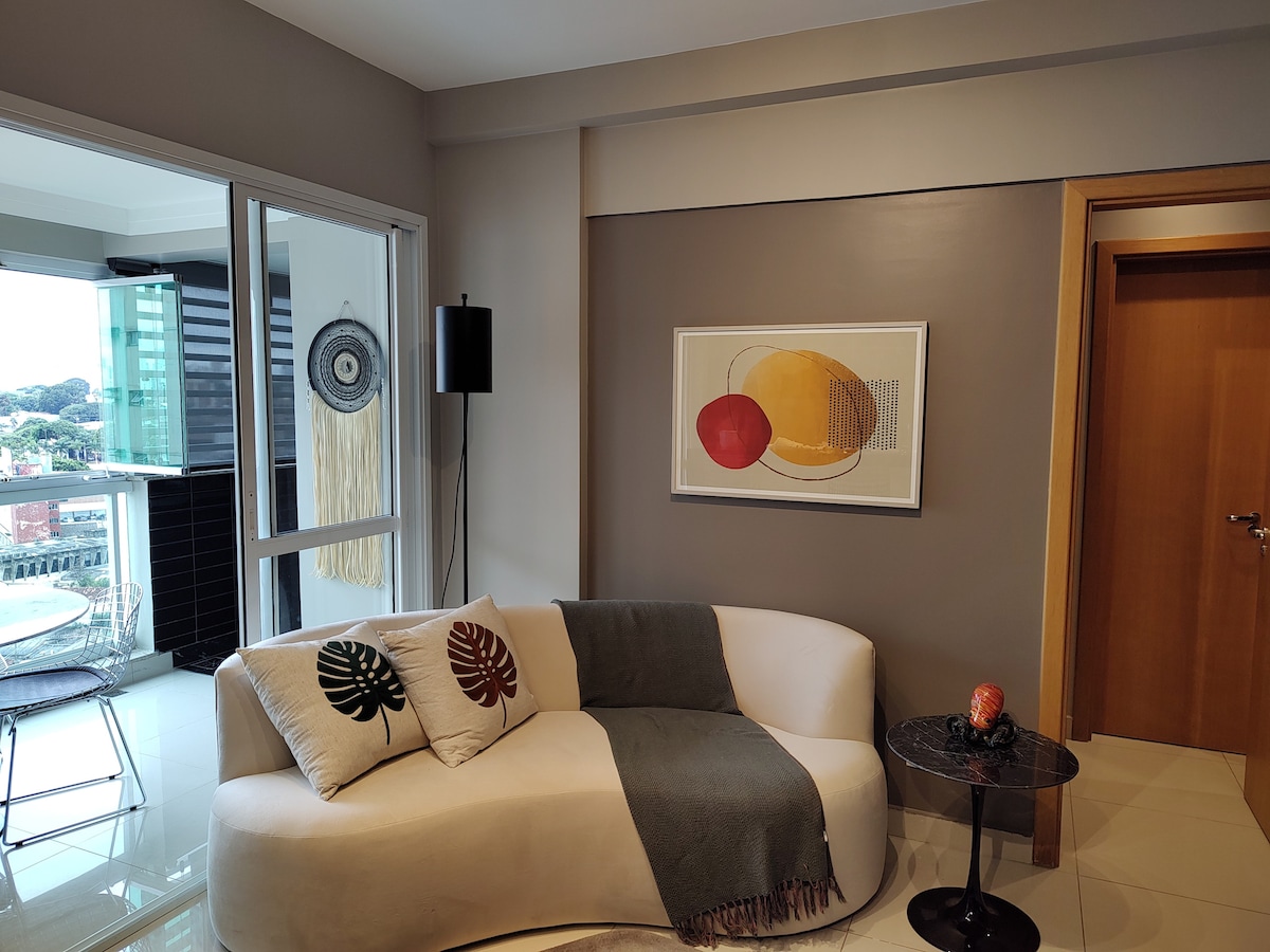 A modern living area is shown, featuring a curved sofa adorned with decorative pillows displaying leaf designs. A small side table holds a sculpture. A colorful abstract artwork hangs on the wall, while large glass doors provide views of the outdoor space.