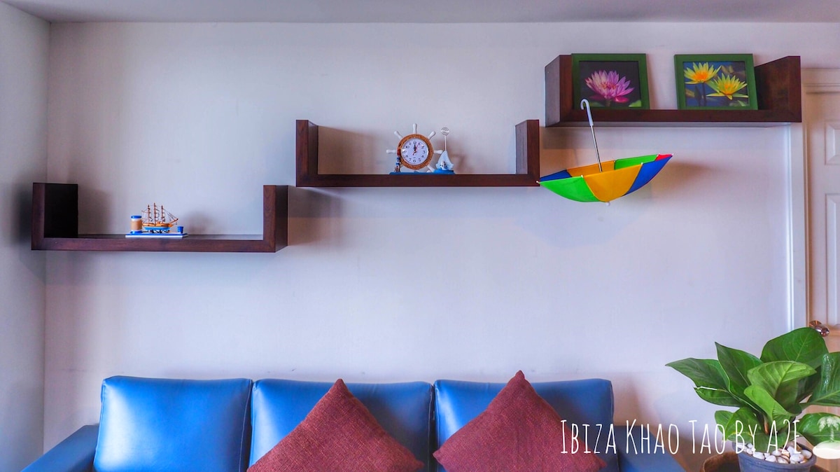 A living area features a blue sofa adorned with two patterned cushions. Wall-mounted wooden shelves display decorative items, including a small clock, an umbrella, and a model ship. A potted plant adds a touch of greenery to the space.