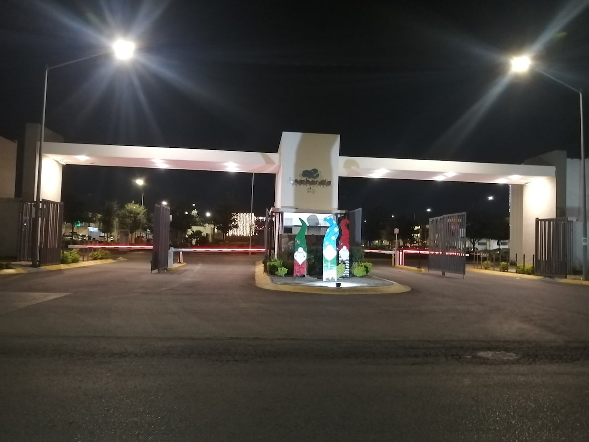 A controlled access entrance is illuminated at night, featuring a sign with a stylized tree emblem. The entrance is flanked by security gates, with soft lighting highlighting the surrounding landscaping. A smooth roadway leads further into the residential area.