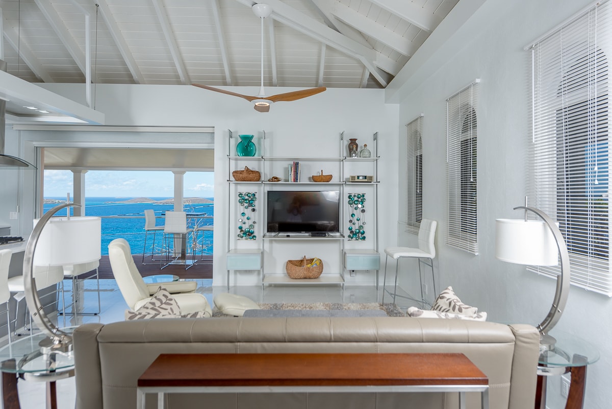 A bright living area is presented with high ceilings and large windows showcasing ocean views. A comfortable sofa is paired with a wooden table, while modern bar stools stand at a counter. Decorative elements can be seen on shelving units, adding a serene touch.
