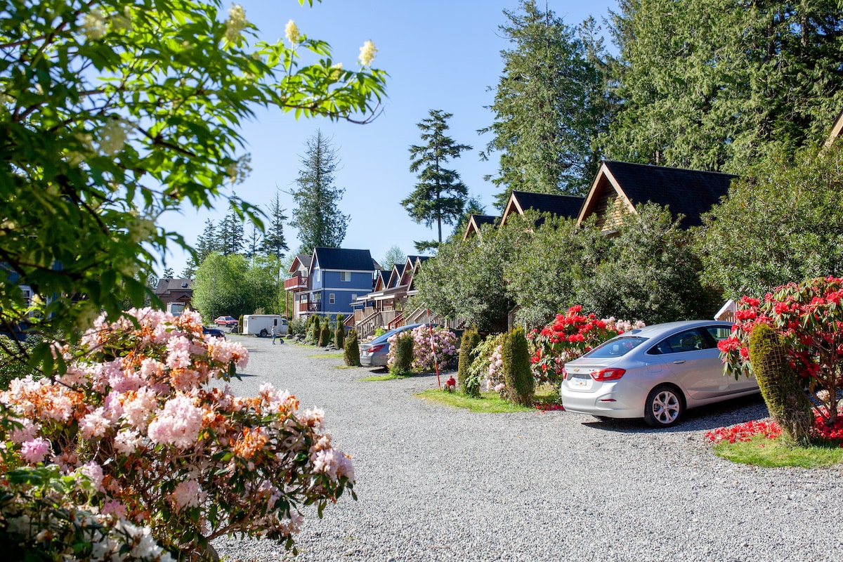A picturesque pathway is lined with vibrant floral blooms, leading to a collection of charming cottage-style buildings. Gravel surfaces provide access to parked vehicles, while lush greenery and trees create a serene atmosphere. The scene captures a warm, inviting environment that is easily accessible.