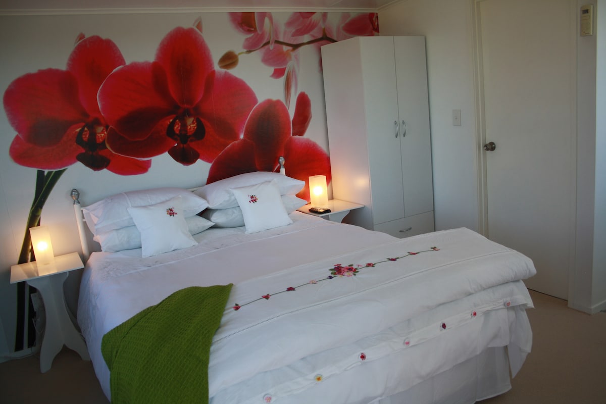 A cozy bedroom features a queen bed adorned with white linens and decorative pillows. Flanking the bed, two nightstands with soft lighting create a serene environment. A floral mural of vibrant red orchids adds a pop of color to the wall, complemented by an airy atmosphere.
