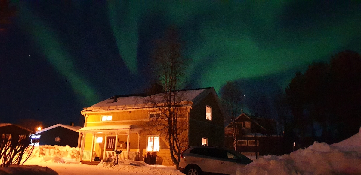 A two-story house is set against a backdrop of a vibrant aurora borealis illuminating the night sky. Snow blankets the ground, while warm light glows from the windows. The surroundings feature additional structures, creating a tranquil winter scene.