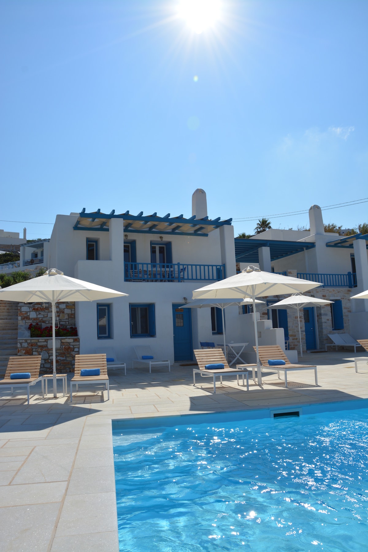 A bright sunny day highlights the elegant design of the accommodation, featuring white structures with blue accents. Lounge chairs surround a clear swimming pool, complemented by shaded umbrellas. The inviting outdoor space emphasizes a relaxing atmosphere for guests.