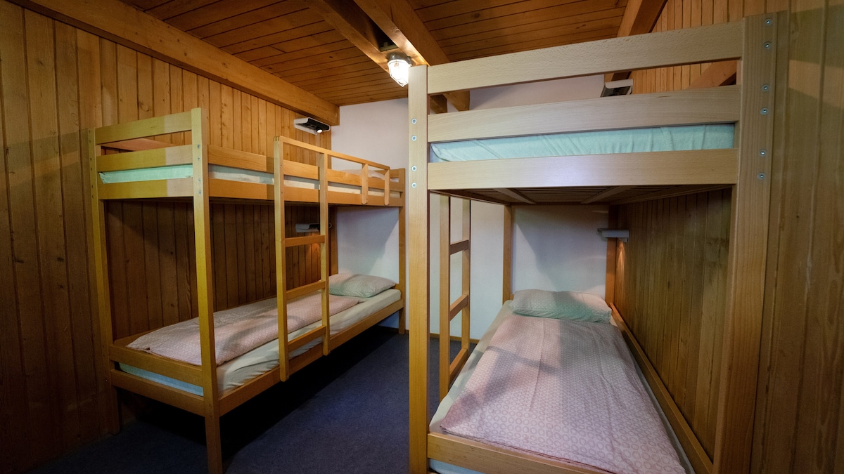 A room features two sets of wooden bunk beds arranged against wooden walls. Soft bedding is provided on each level, with individual reading lights attached to the bed frames. A warm atmosphere is complemented by overhead light fixtures and simple blue carpeting.