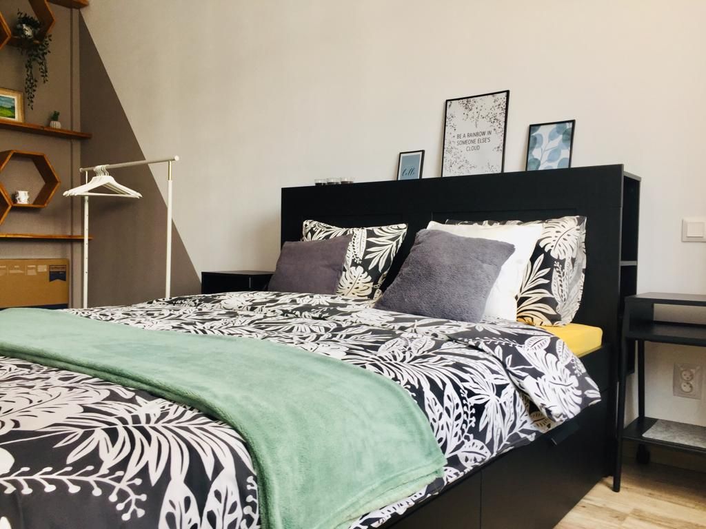 A spacious bed is dressed in a floral-patterned duvet cover, complemented by gray pillows. A soft green throw blanket adds a touch of color. The headboard features functional shelving with framed art pieces, and a clothing rack is positioned nearby.