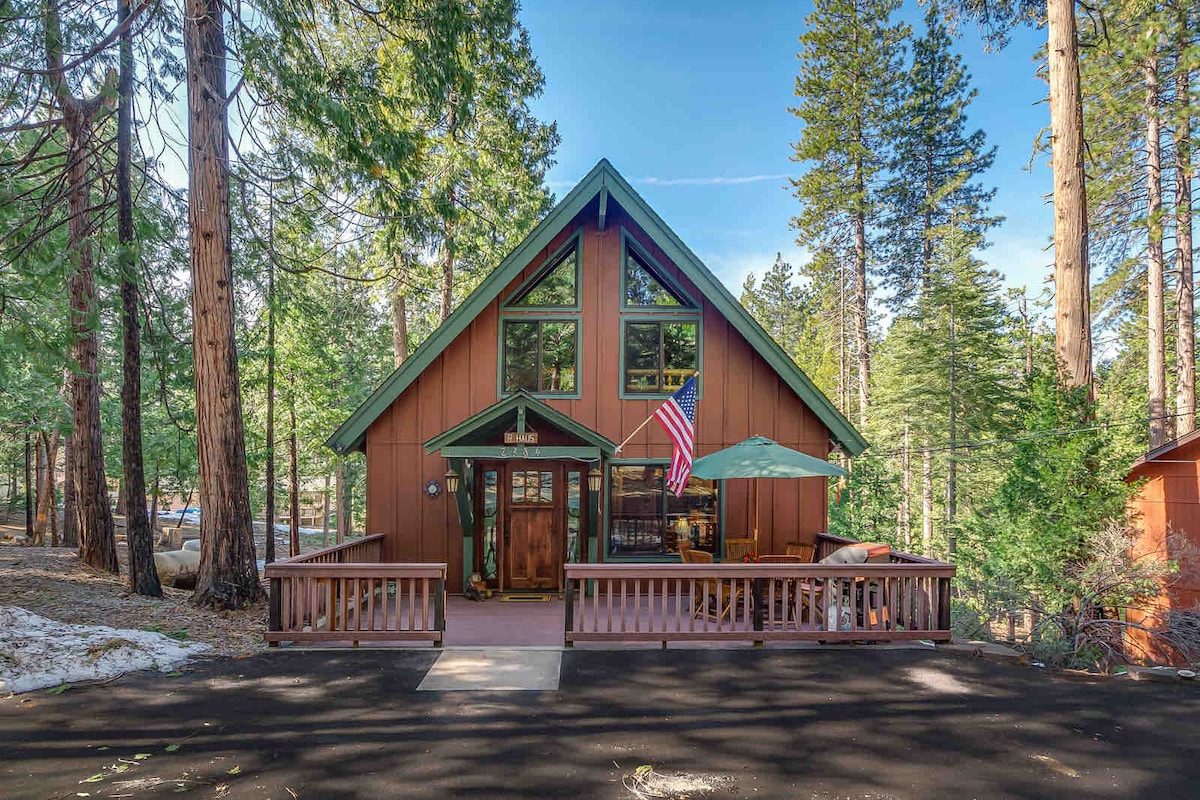 The cabin's exterior features awooden facade with a sloped roof and large windows, surrounded by tall pine trees. A welcoming front porch is visible, equipped with a table and umbrella. A flag hangs proudly from the front, displaying a sense of home.