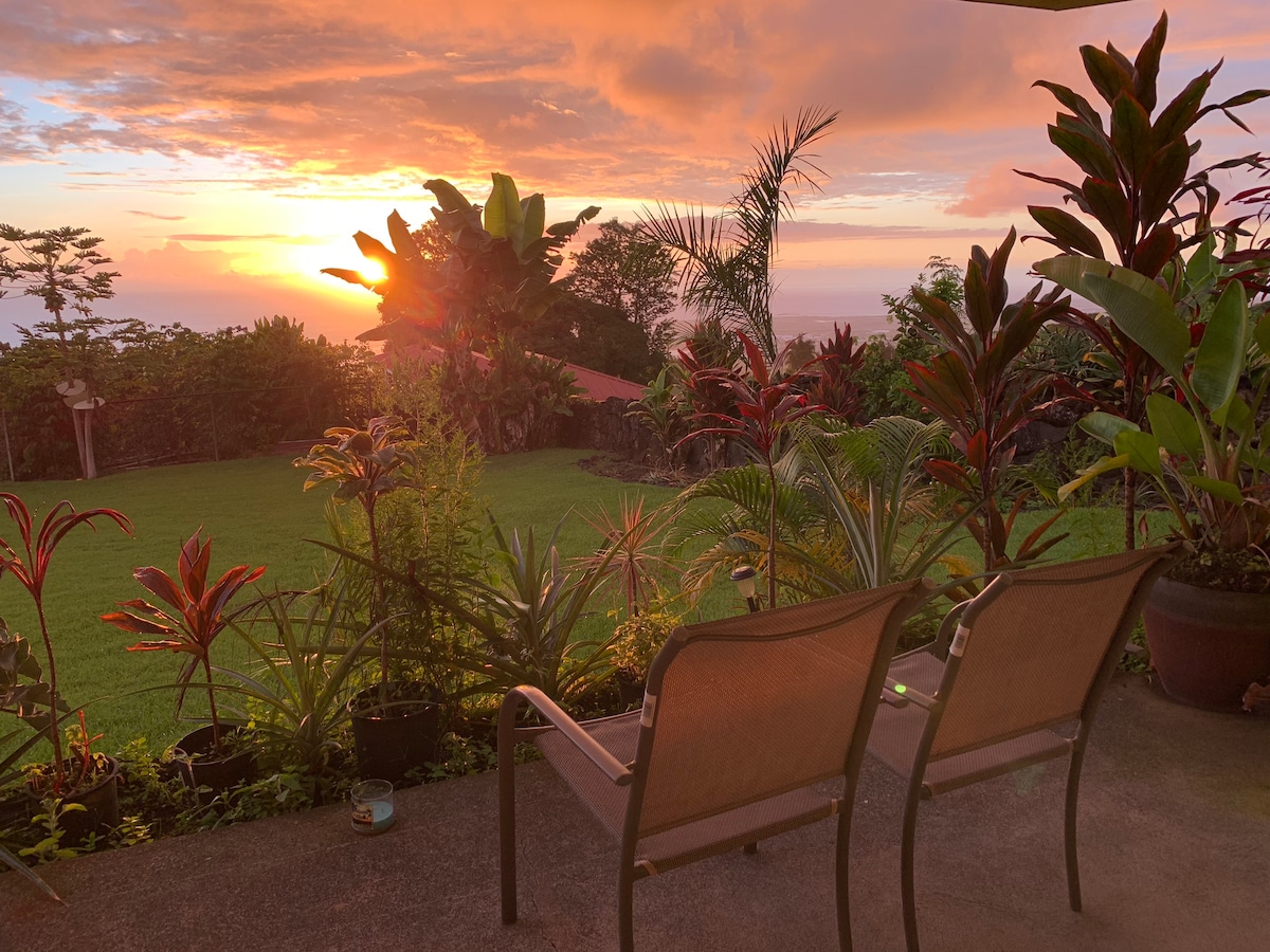 A covered patio showcases two comfortable lounge chairs facing a vibrant sunset over the ocean. Lush tropical plants and trees surround the area, creating a serene atmosphere. The expansive grassy yard extends beyond, providing a perfect setting for enjoying nature and the evening sky.
