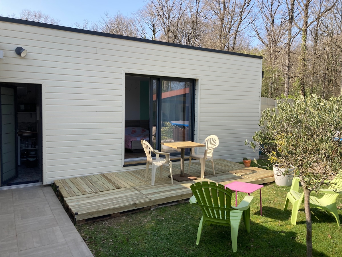 An outdoor space features a wooden deck with a small table and two chairs. Bright green lounge chairs are positioned nearby on the lawn. Large glass doors provide access to the studio interior, surrounded by trees that provide a natural setting.