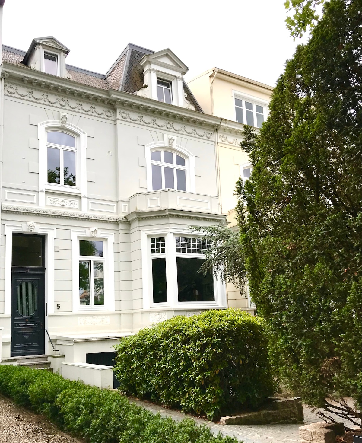 The exterior of a charming city villa features a classic facade with large windows. A well-maintained garden surrounds the entrance, showcasing lush greenery and carefully trimmed bushes. The welcoming entrance is complemented by a contrasting dark door, enhancing the overall architectural harmony.