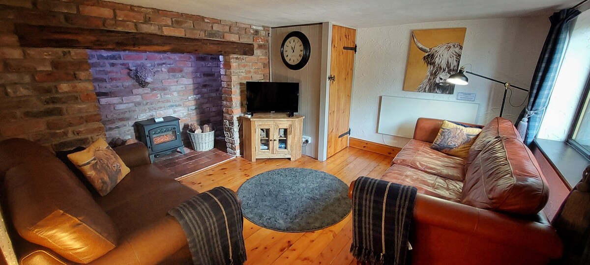 A cozy living area features a combination of brown leather sofas arranged around a circular area rug. A wood-burning stove is set into a brick wall, complementing the rustic decor highlighted by a round wall clock and artwork of a Highland cow.