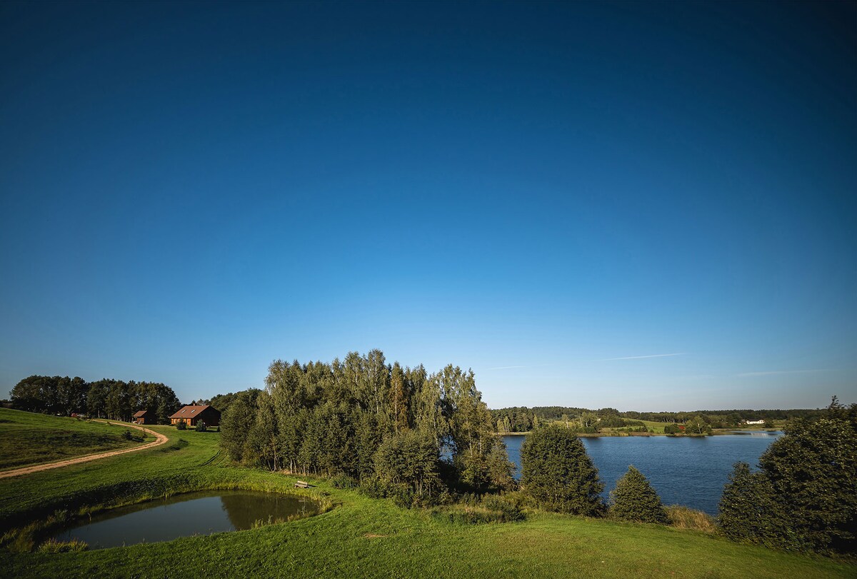 A scenic view of the Raudeņu lake is captured, bordered by lush green fields and trees. A dirt path winds through the landscape, leading to traditional wooden houses in the distance. The clear blue sky enhances the tranquil setting.