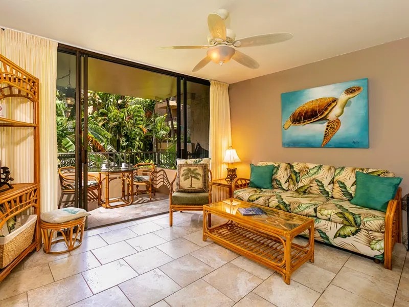 The living area features a rattan furniture set, including a sofa adorned with tropical print cushions. A glass coffee table rests on tiled flooring, while large glass doors provide views of the lush garden outside. A ceiling fan adds to the open, airy ambiance.