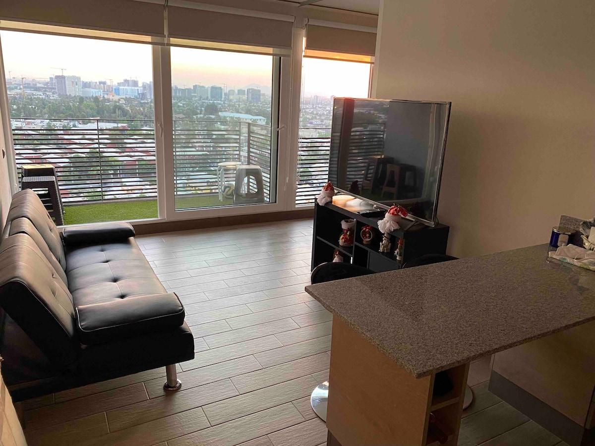A modern living space features a sleek black sofa facing a large flat-screen television. Floor-to-ceiling windows provide views of the city skyline, with soft natural light flooding the room. A kitchen island with a stone countertop is visible, enhancing the inviting area.