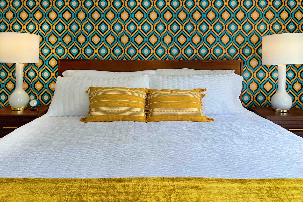 A spacious bed is adorned with a white quilt and decorative pillows in shades of yellow. The backdrop features a vibrant mid-century modern wallpaper with a geometric pattern in teal and gold. Soft lighting is provided by bedside lamps on each side.