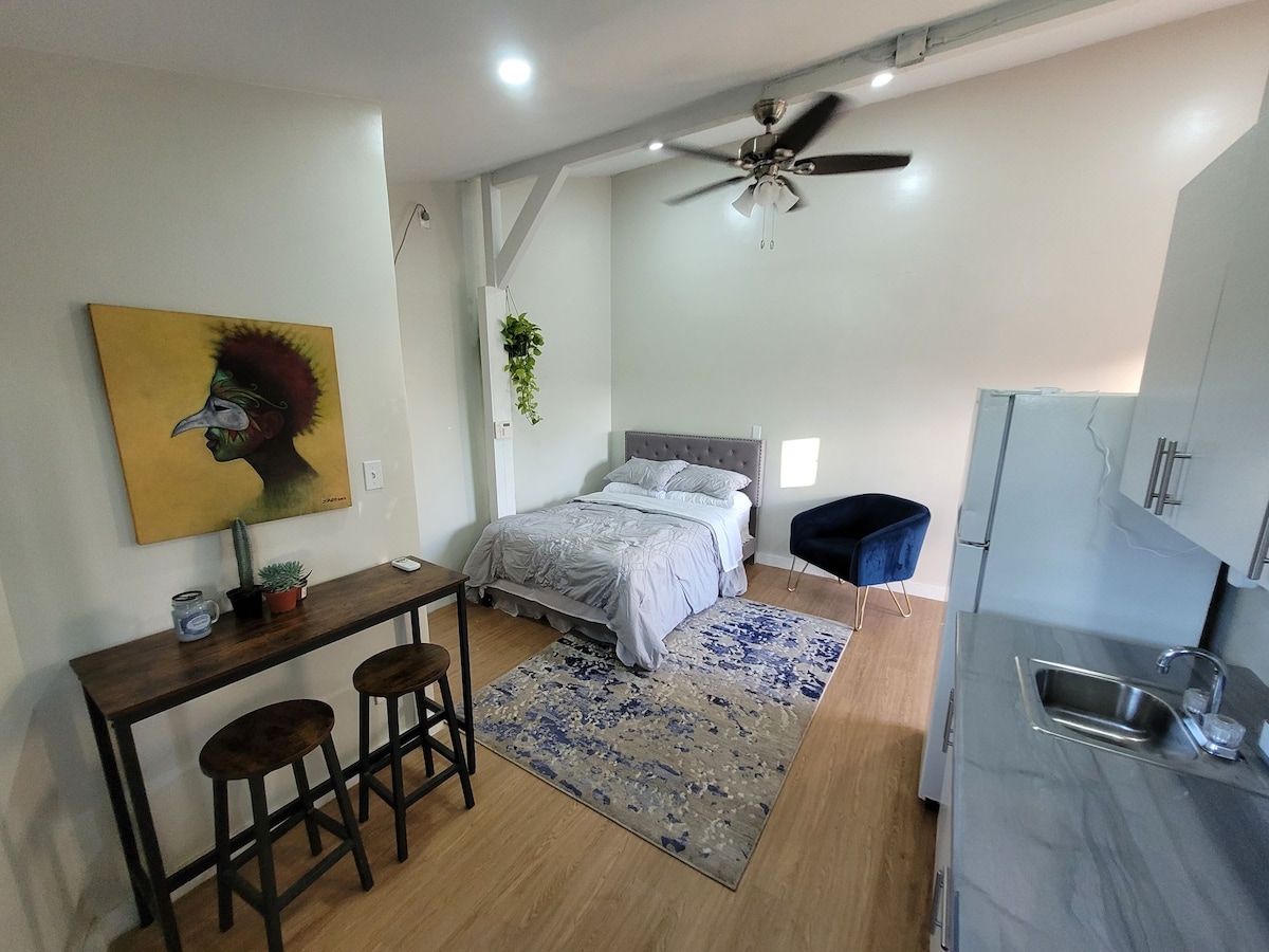 The cozy studio space features a comfortable bed arranged with soft bedding. A small table and two stools are positioned nearby. A blue accent chair adds a pop of color, while a decorative rug complements the minimalist design. Natural light enters through a window.