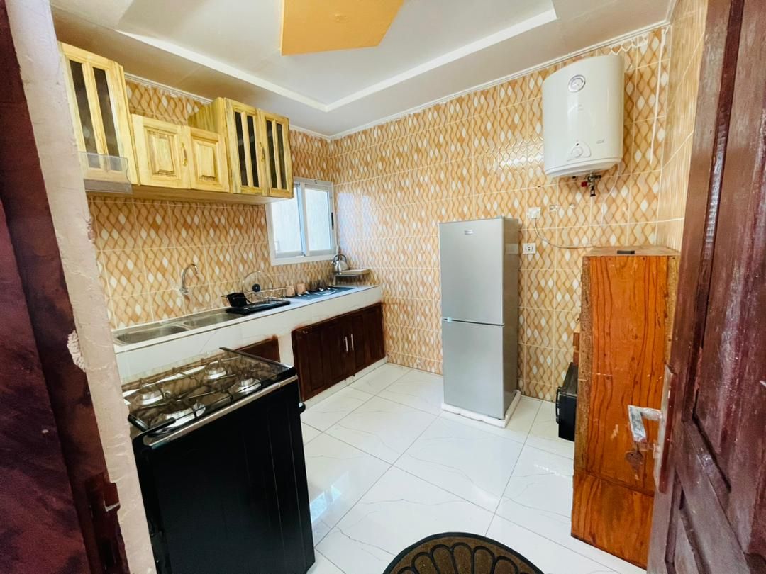 The kitchen features wooden cabinetry and a modern refrigerator. A stove is positioned beside a large sink, with ample counter space available for meal preparation. Tile flooring enhances the room's brightness, complemented by a window that allows natural light to enter.