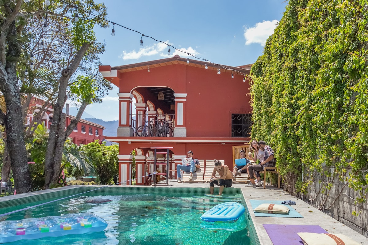 A vibrant pool area features a clear water surface surrounded by lush greenery and a well-maintained terrace. A handsome red building stands in the background, with its distinctive architecture highlighted by the bright sunlight. Lounge chairs and pool accessories are arranged neatly by the water.