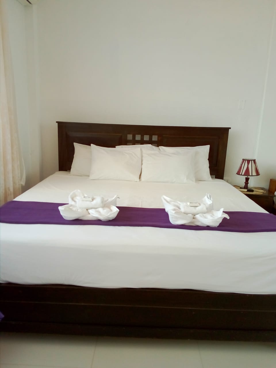 A spacious bed is centered in the room, adorned with neatly arranged white pillows. A lavender runner adds a pop of color across the white bedding. A bedside table with a lamp complements the serene atmosphere, while natural light filters in through a nearby window covered with sheer curtains.