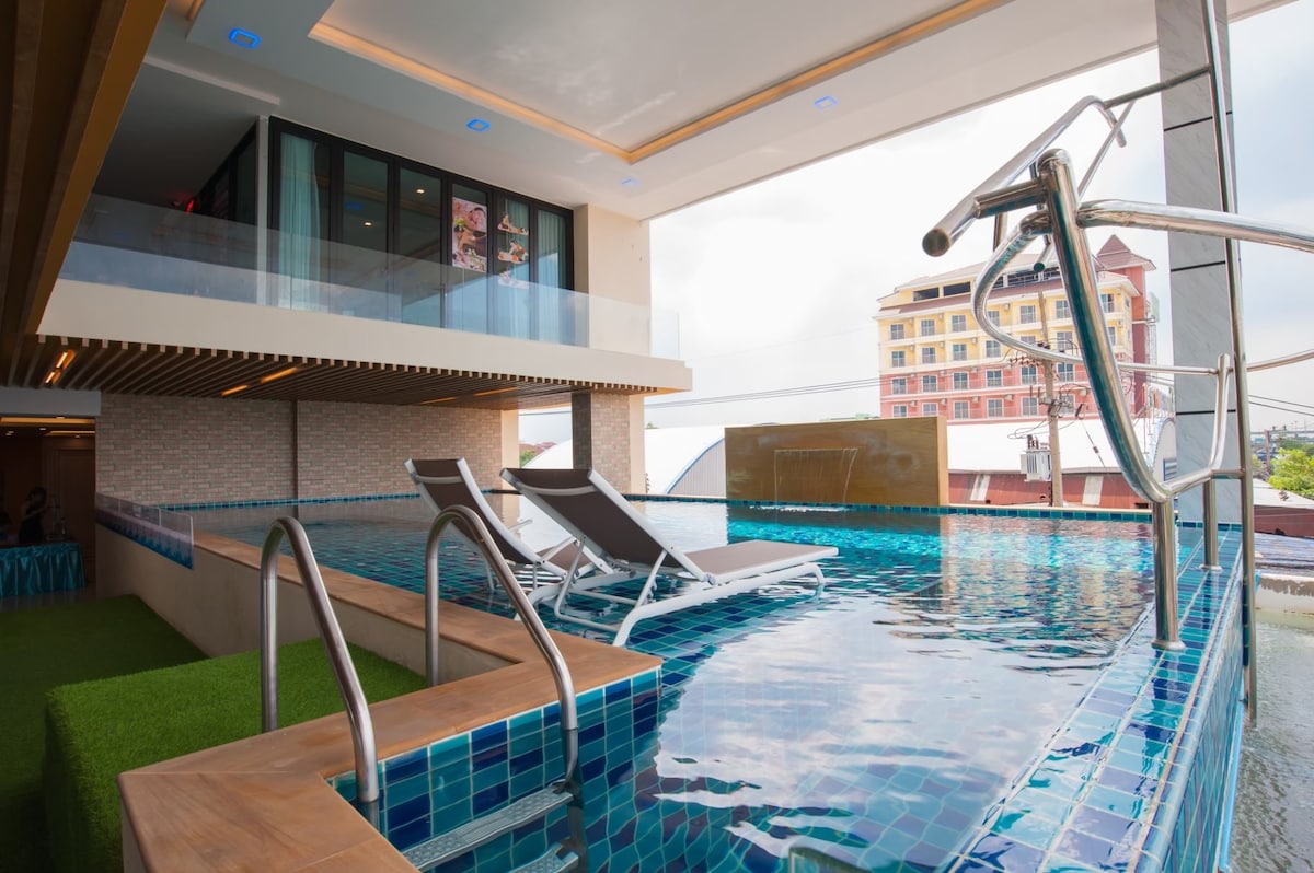 A modern rooftop pool area is depicted, featuring two white lounge chairs positioned along the edge. The pool's turquoise tiles reflect the surrounding skyline, while glass railings provide an unobstructed view. Lush greenery is seen adjacent to the pool, enhancing the serene environment.