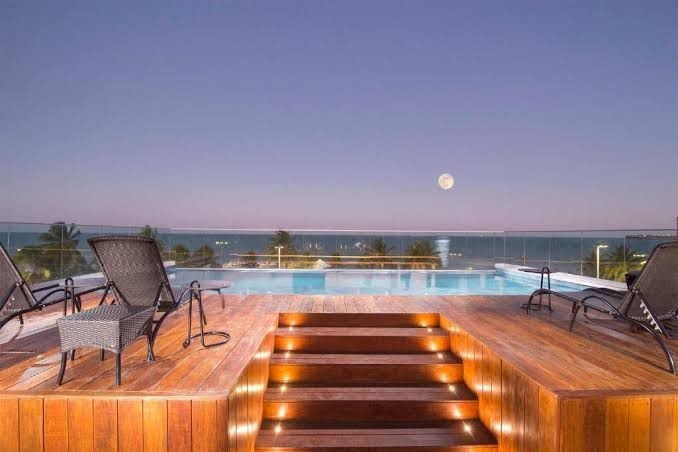 A tranquil pool area features wooden decking leading to a serene infinity pool, with several loungers arranged for relaxation. The expansive view includes the ocean and a clear sky, where a full moon illuminates the scene at dusk.