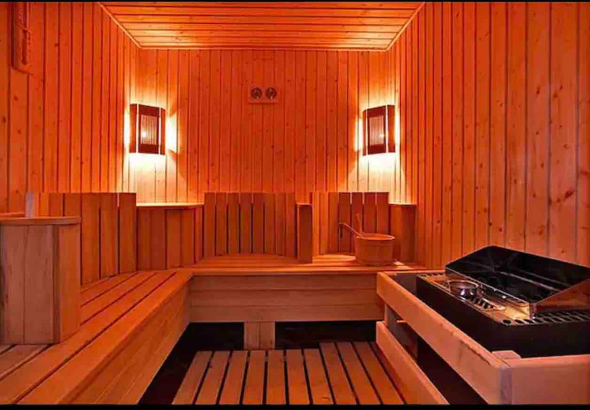A wooden sauna room is designed with warm lighting fixtures mounted on the walls. Benches made of slatted wood create seating arrangements along the walls. In one corner, a traditional sauna stove is visible, accompanied by a wooden bucket and ladle.